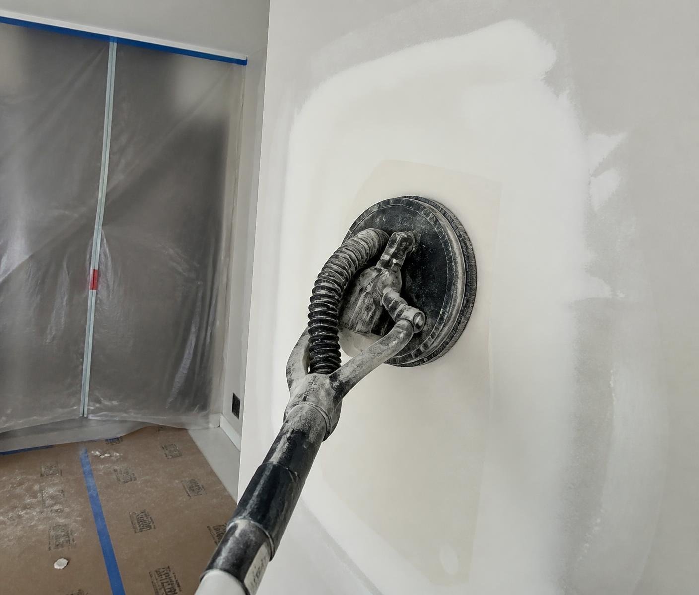 Pole sander smoothing a drywall patch with plastic containment and floor protection